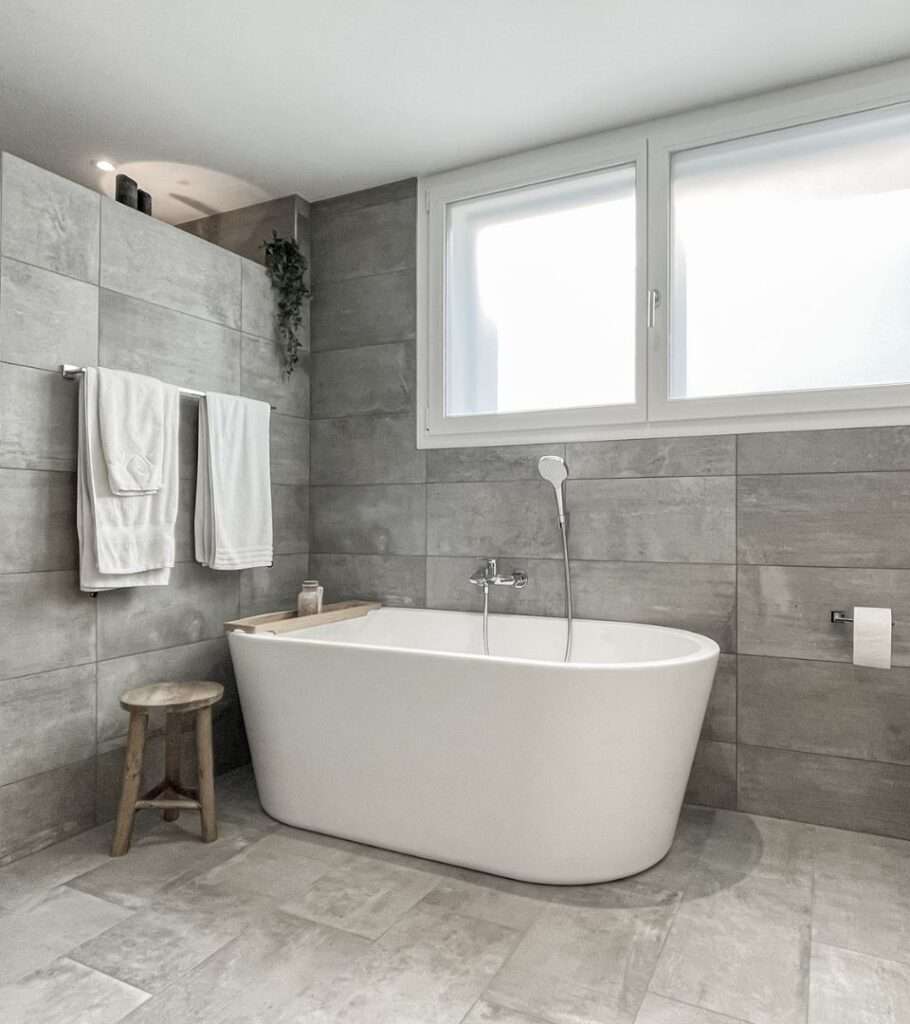 Modernes Badezimmer mit freistehender Badewanne, grauen Fliesen und großem Fenster