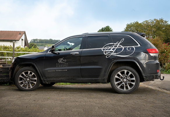Auto mit Aufschrift von Distillerie Zimmerli auf einem Platz mit Bäumen im Hintergrund